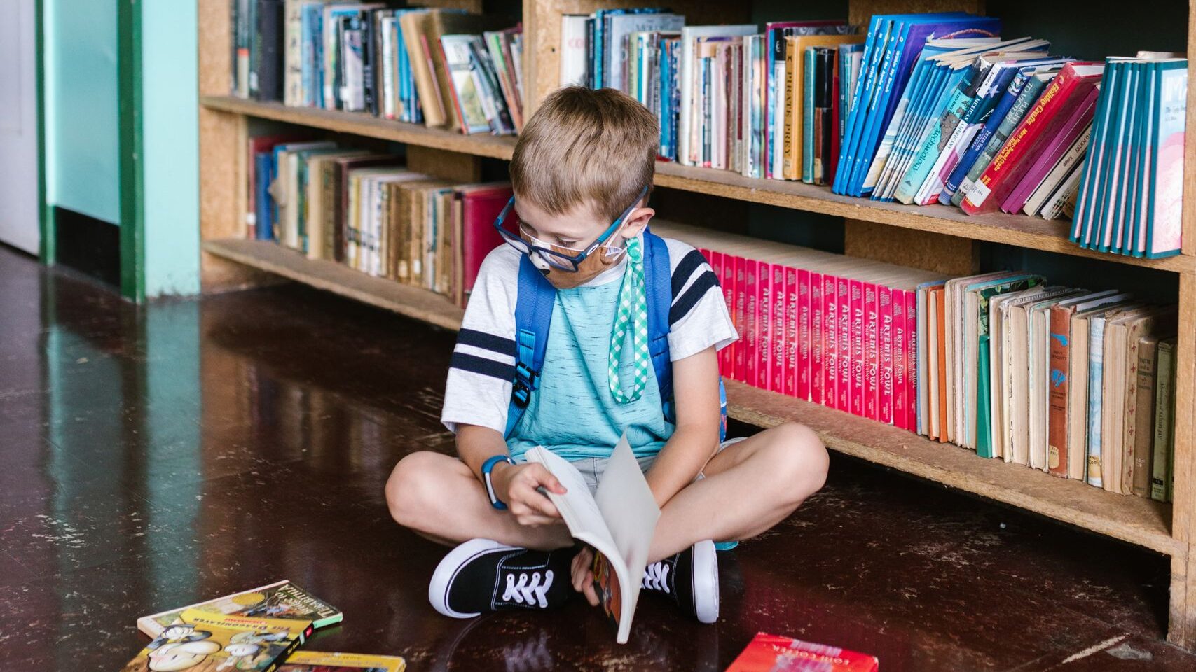 A child enjoys reading books inside a library, surrounded by shelves.