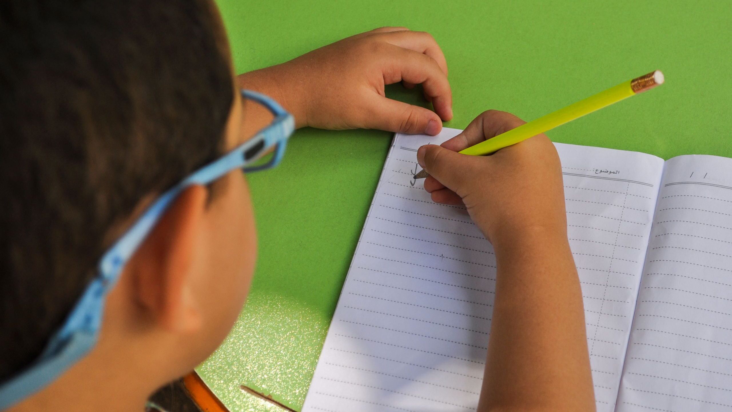 A child wearing glasses writes in a lined notebook with a yellow pencil on a green table.