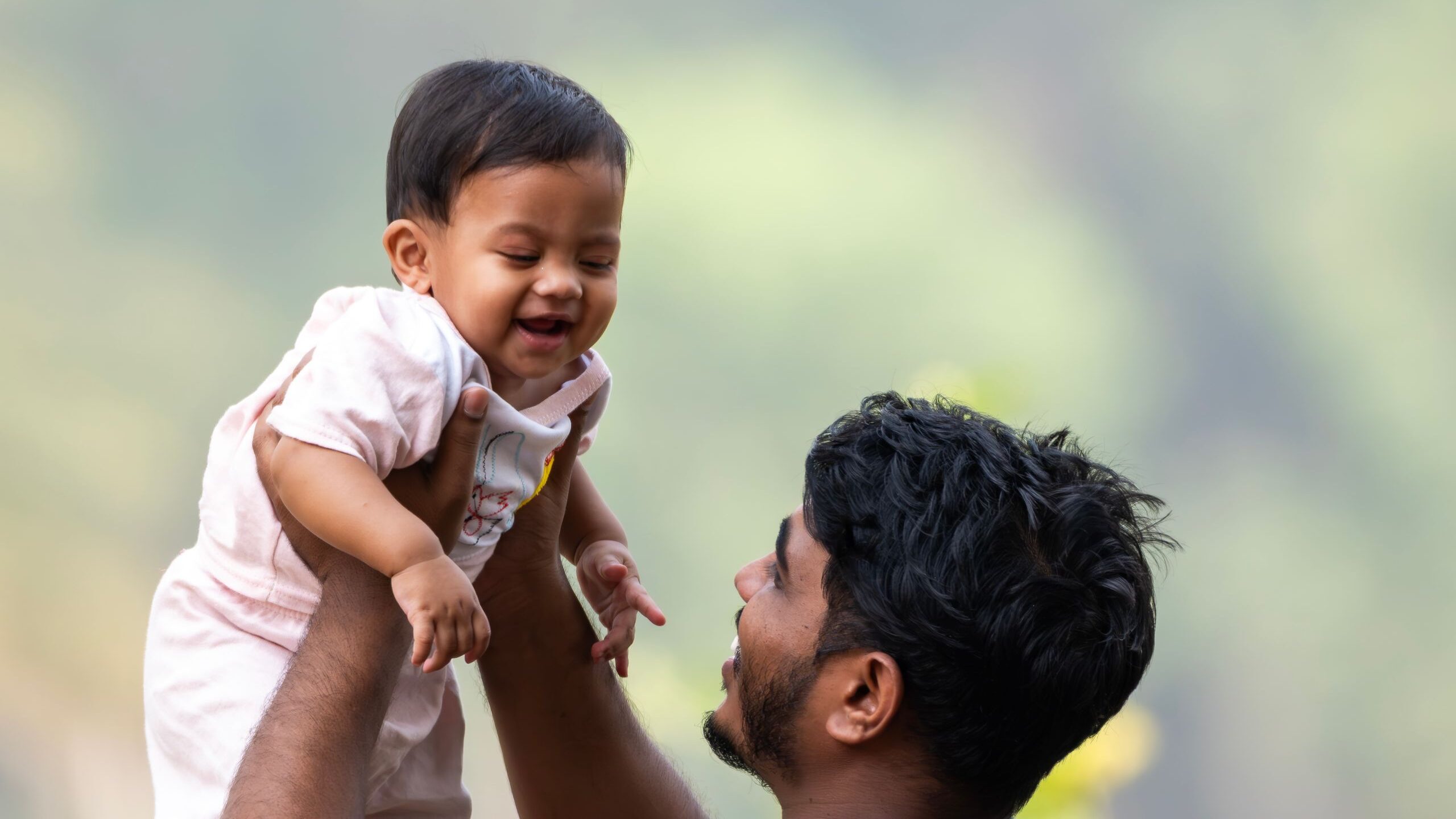 A father joyfully lifts his smiling baby in an outdoor setting, capturing a moment of pure happiness.