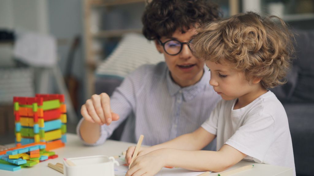 A mother and son engage in a creative drawing session at a table indoors, surrounded by colorful blocks.