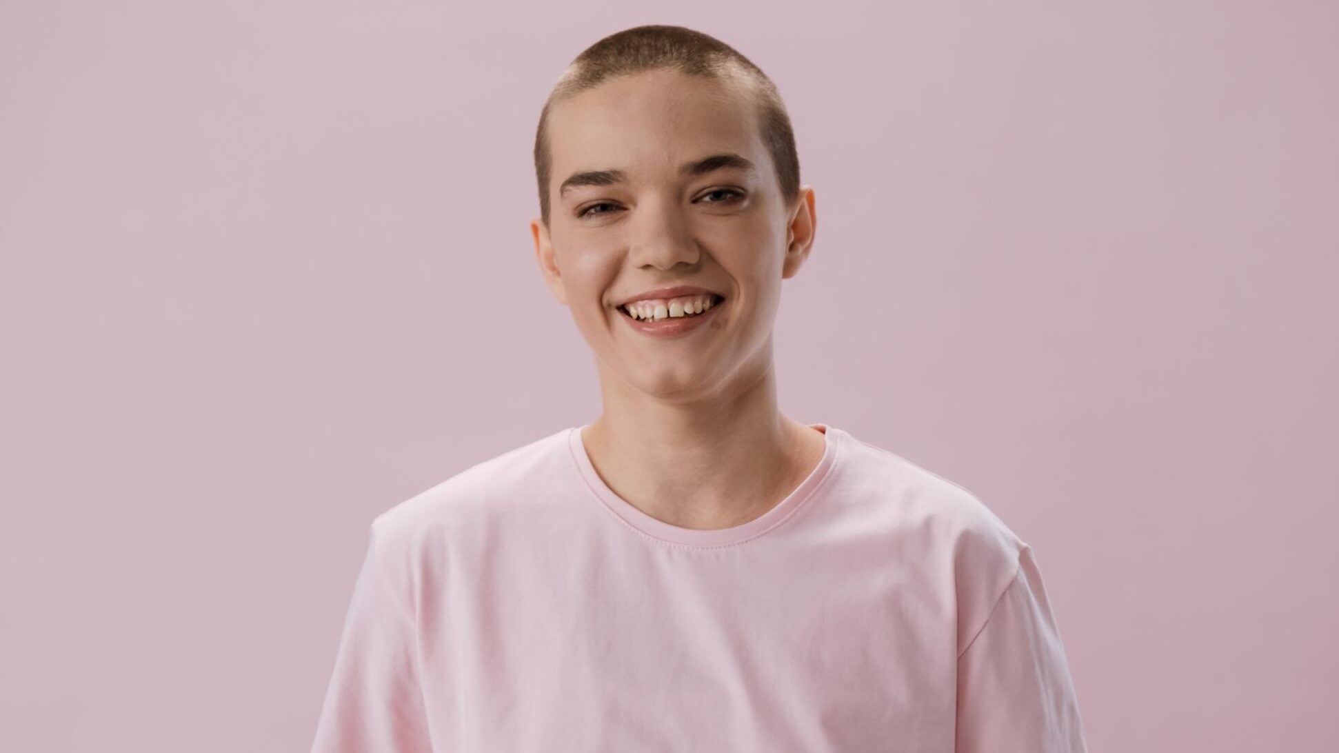 A smiling woman with a shaved head wears a pink shirt symbolizing breast cancer awareness.