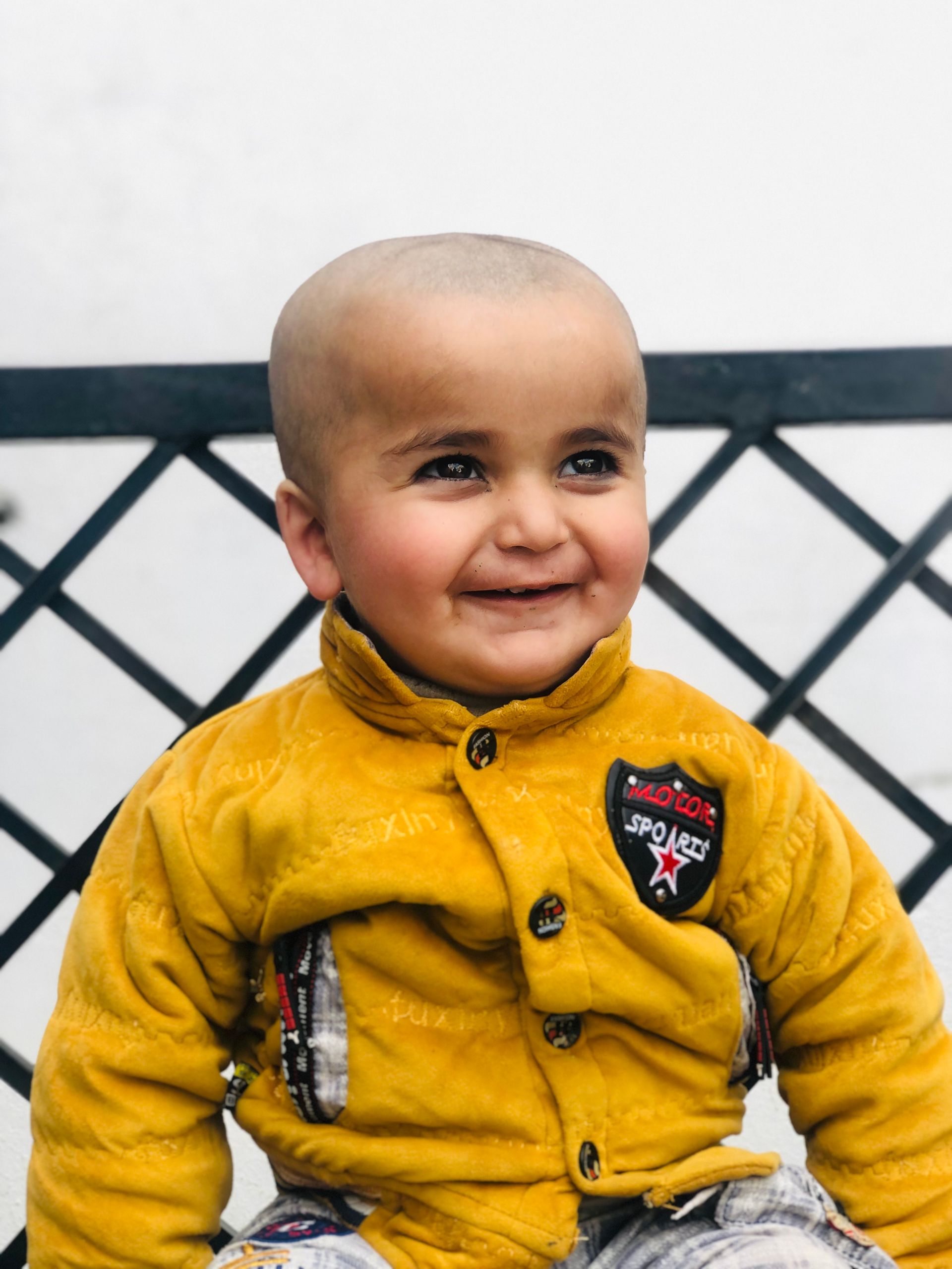 Cute baby with a shaved head in yellow jacket smiling outdoors, portraying joy and innocence.